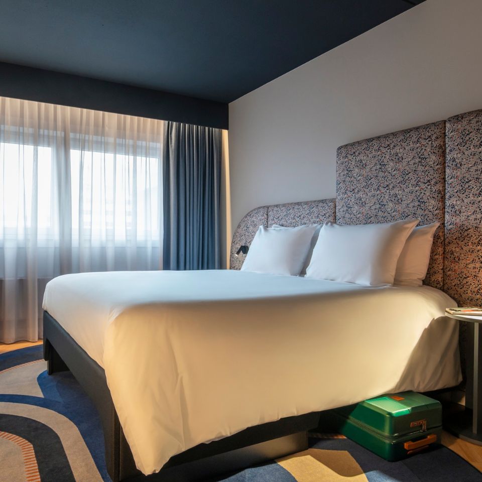 A modern hotel room with a plush bed, white linens, and a patterned headboard. Sunlight filters through sheer curtains, creating a calm, inviting atmosphere.
