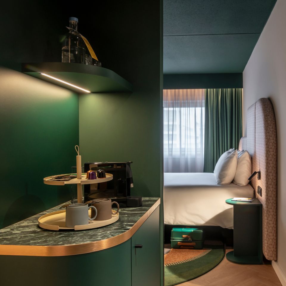 Modern hotel room with green and beige tones. A coffee station with mugs and a machine is in the foreground, and a neatly made bed near a window in the background.