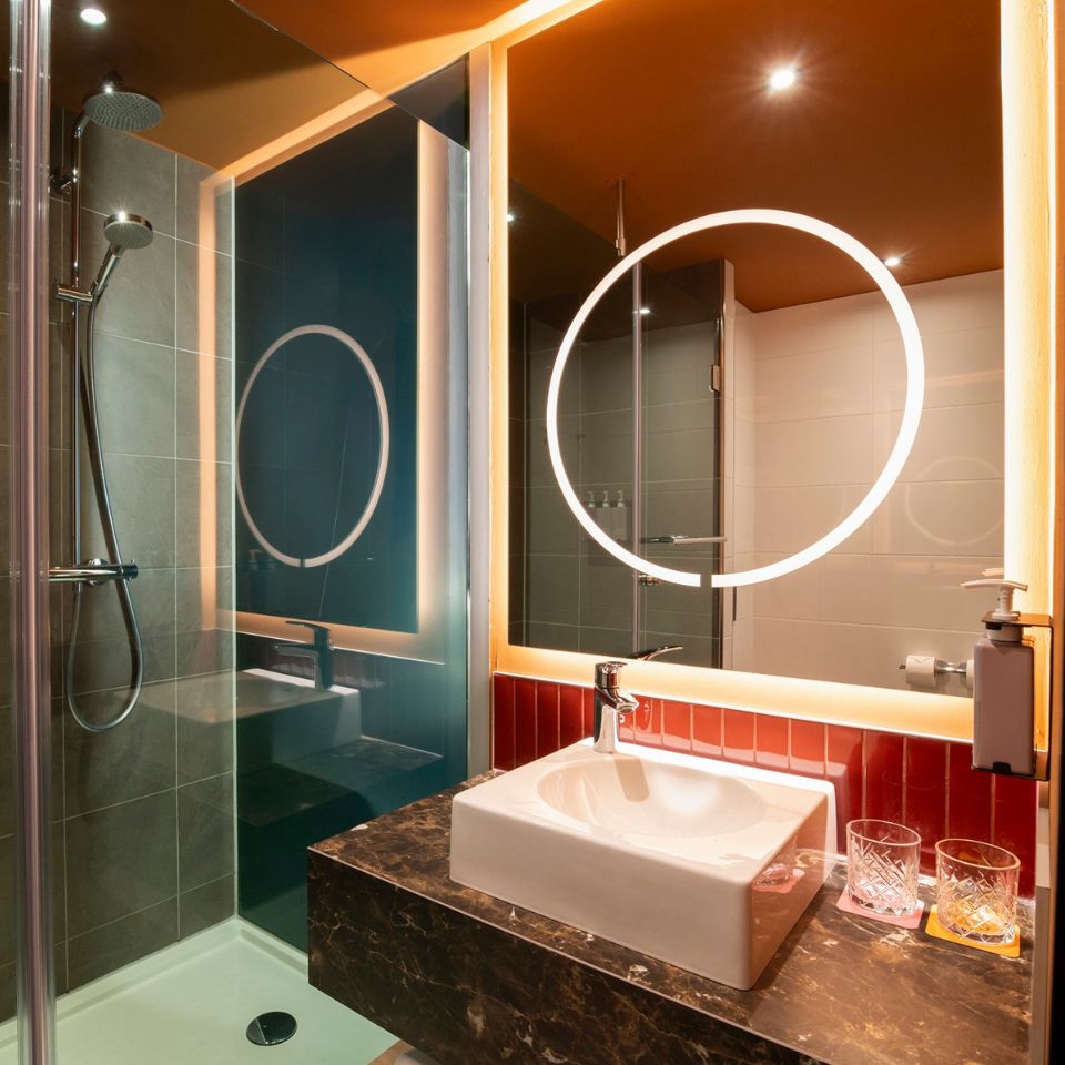 Modern bathroom with warm lighting. Features include a round backlit mirror, a sleek shower, a white sink on a marble counter, and red tiled accents.
