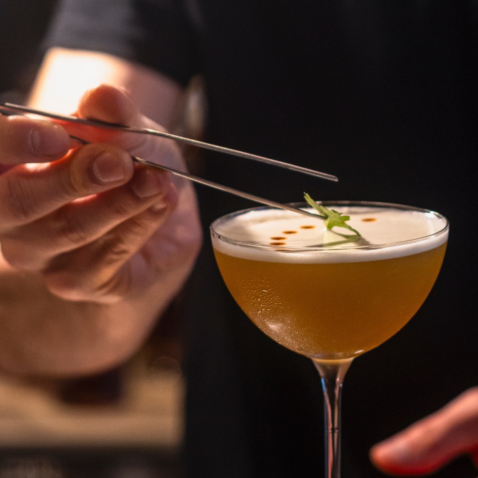 A hand uses tweezers to place a garnish on a frothy, amber cocktail in an elegant glass. The setting is warm and cozy, emphasizing precision and elegance.