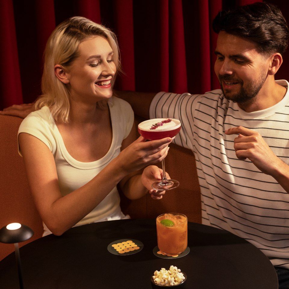 A smiling woman holds a red cocktail while sitting beside a man in a striped shirt. They are at a table with popcorn and an orange drink, against red curtains.