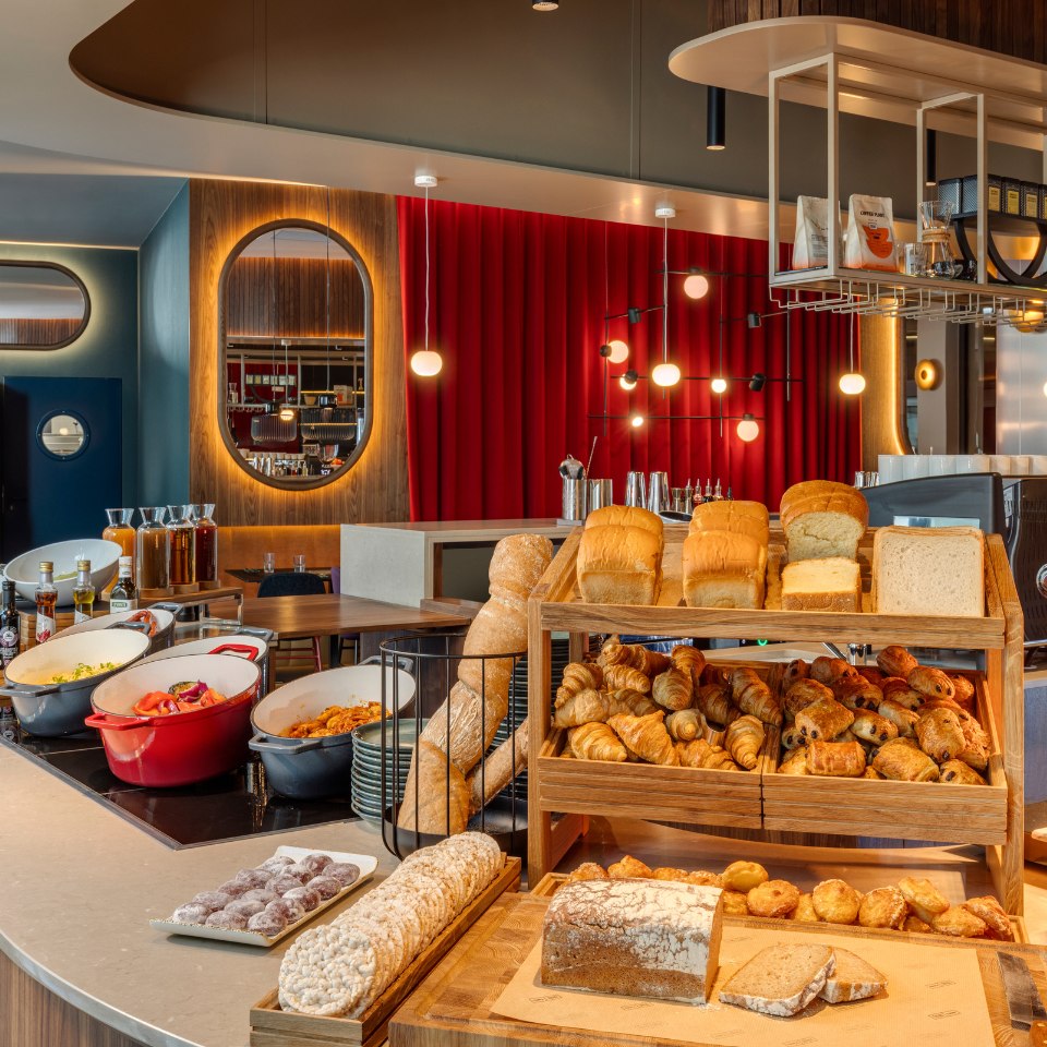 Wooden shelves display various breads and pastries, including croissants and loaves, in a warm, inviting cafe setting with red curtains and modern lighting.