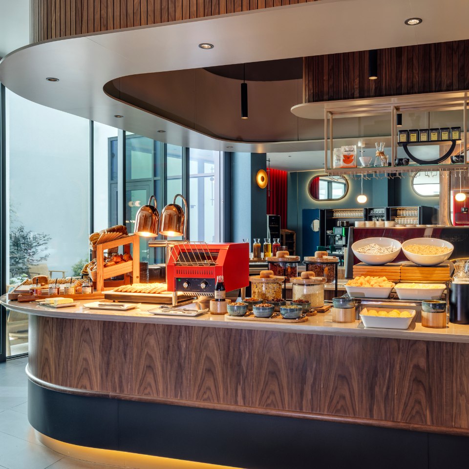 Modern breakfast buffet display with cereals, pastries, and bowls of food on a curved wooden counter. Warm lighting creates an inviting atmosphere.
