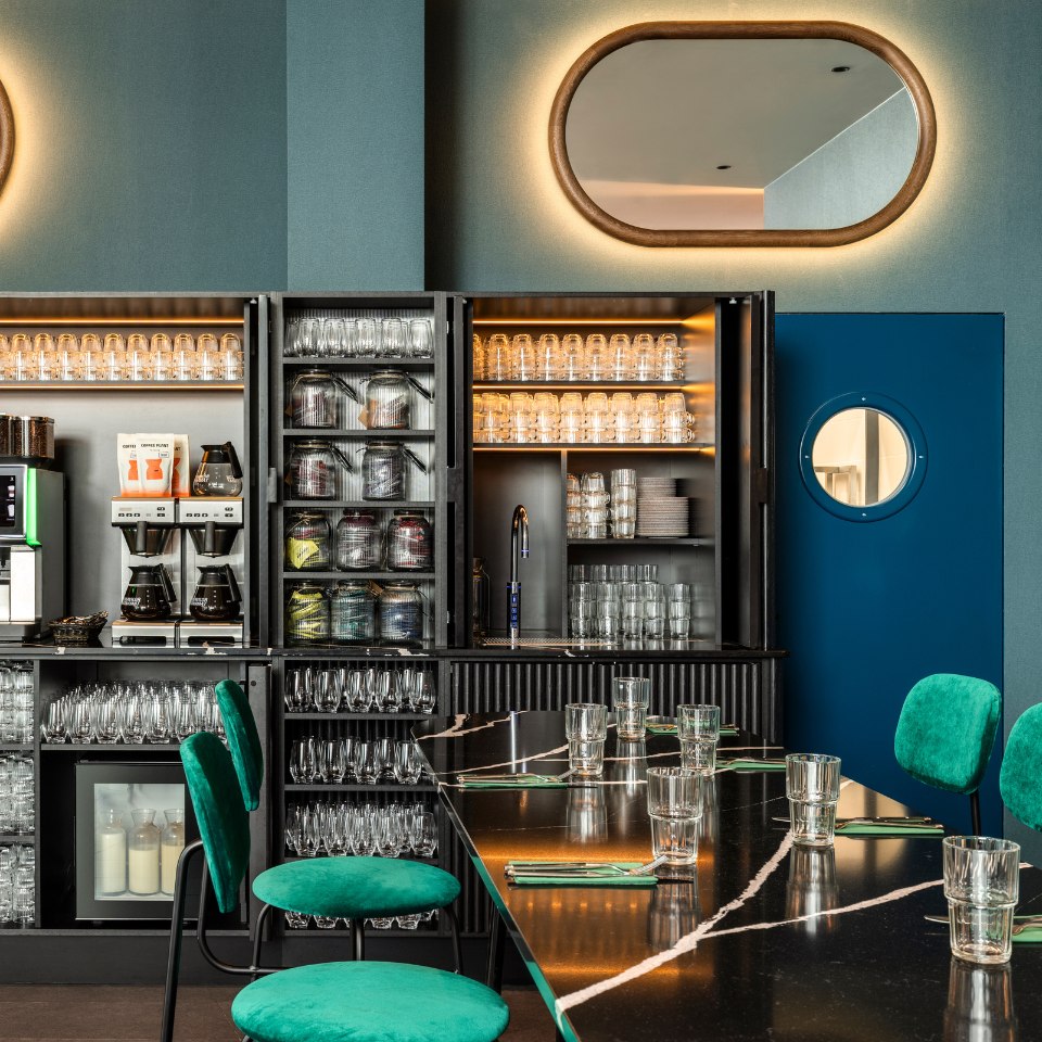 Chic bar interior with a dark counter set with glasses, emerald green chairs, illuminated mirrors, and shelves of glassware. Modern and inviting ambiance.