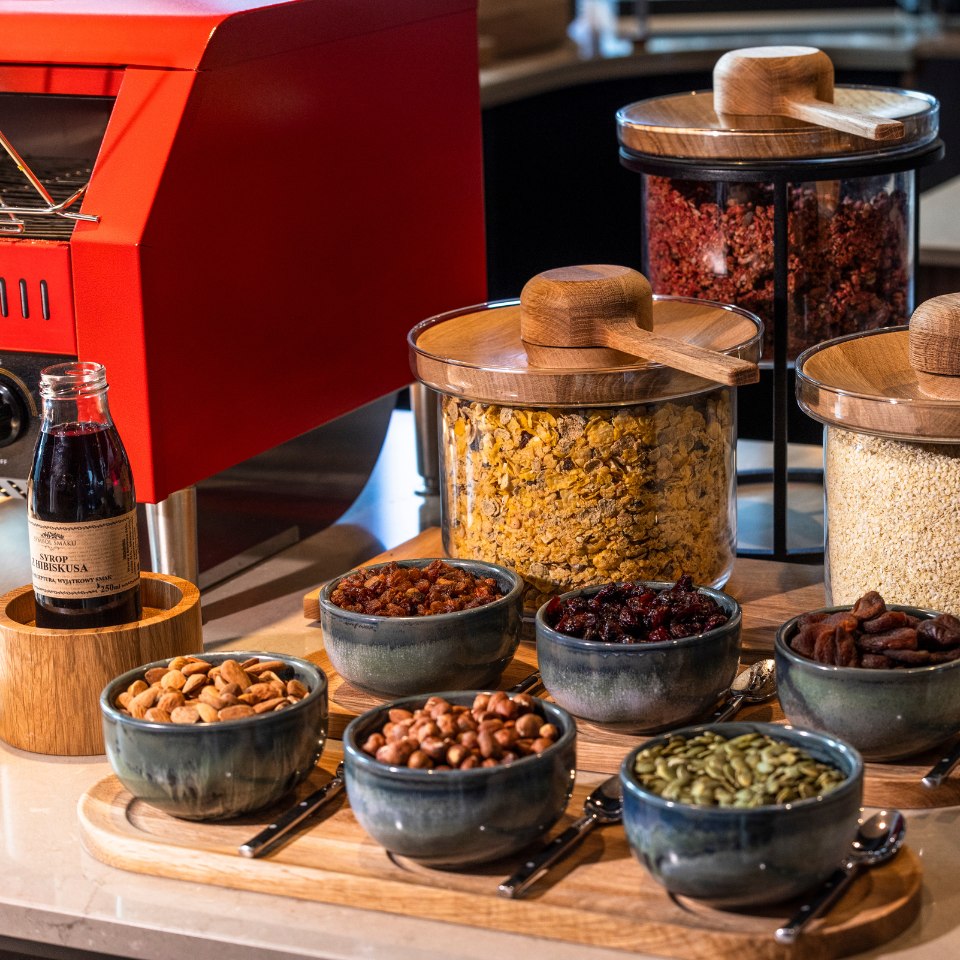 Assorted breakfast items on display include cereals, nuts, dried fruits, and seeds in bowls, with a red toaster and bottled syrup. Warm, inviting setup.