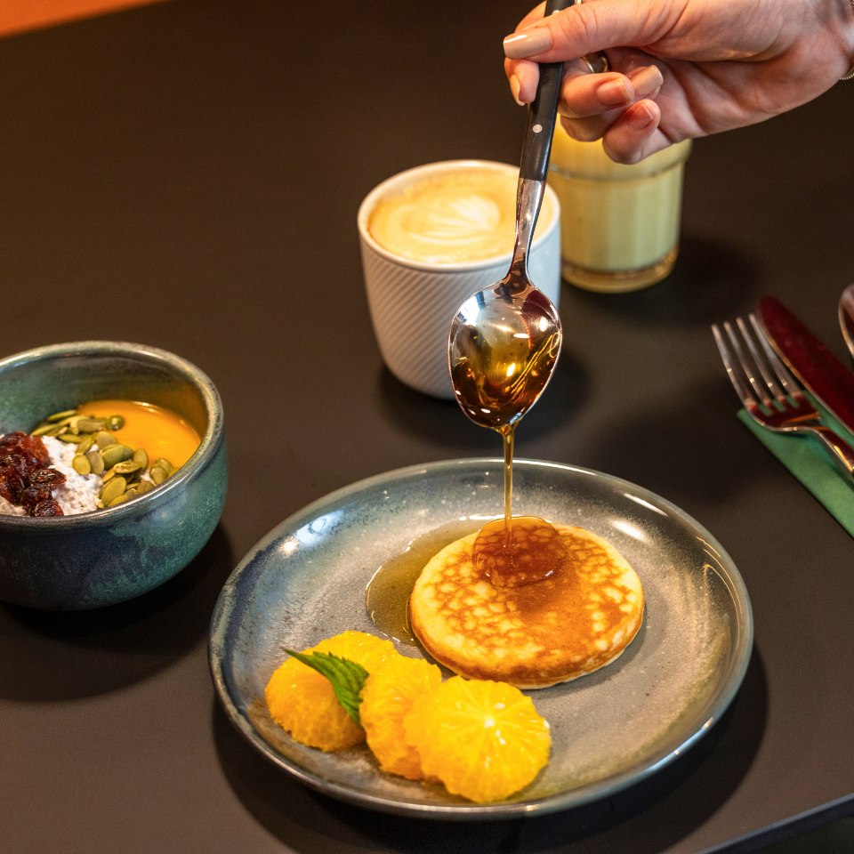A hand drizzles syrup onto a pancake on a blue plate with orange slices. Nearby are a coffee cup and bowl of granola, creating a cozy breakfast scene.