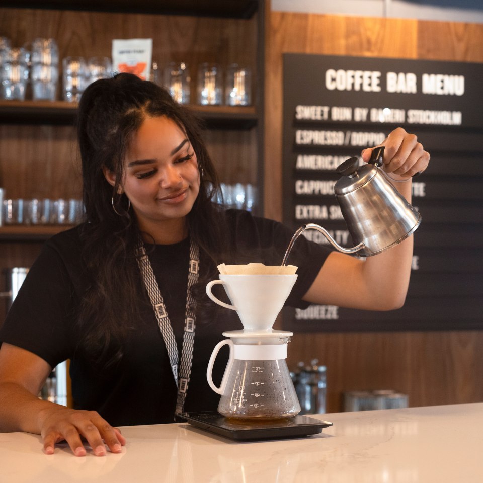 A woman pours hot water into a pour-over coffee maker at a cafe bar. She smiles gently, creating a warm, inviting atmosphere.