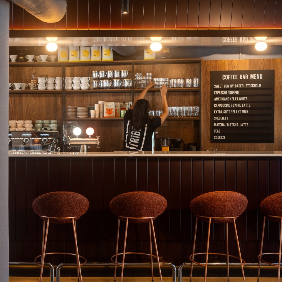 A barista reaches for a glass on a wooden shelf in a cozy coffee shop. The warm lighting highlights a modern, rustic interior with a menu board.