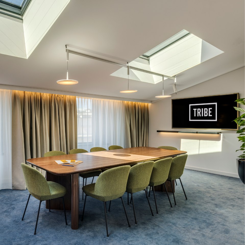 Modern conference room with a long wooden table, green chairs, and a "TRIBE" logo on the wall. Skylights and curtains create a bright ambiance. Calm tone.