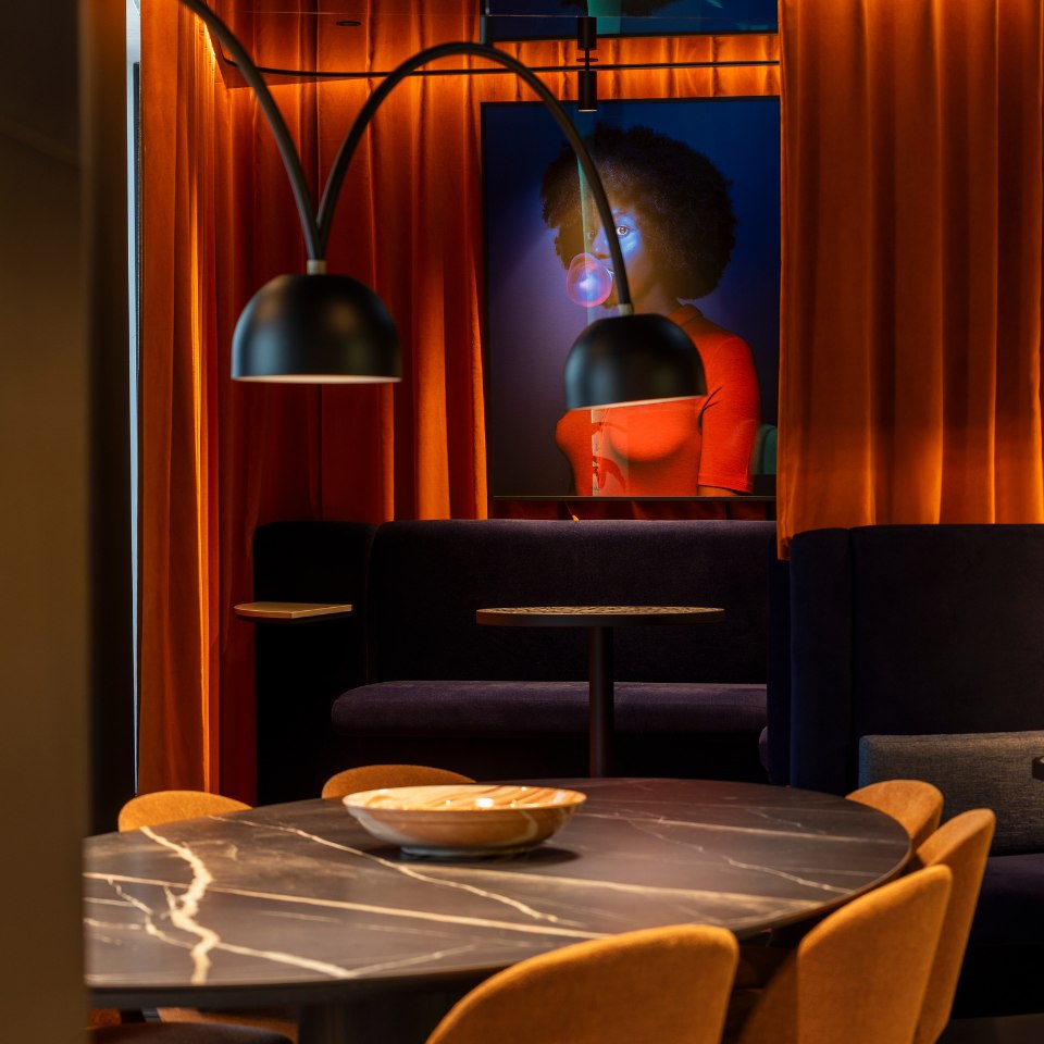 Cozy interior with a round marble table and orange chairs set against rich orange drapes. A painting of a person blowing bubblegum adds a playful touch.