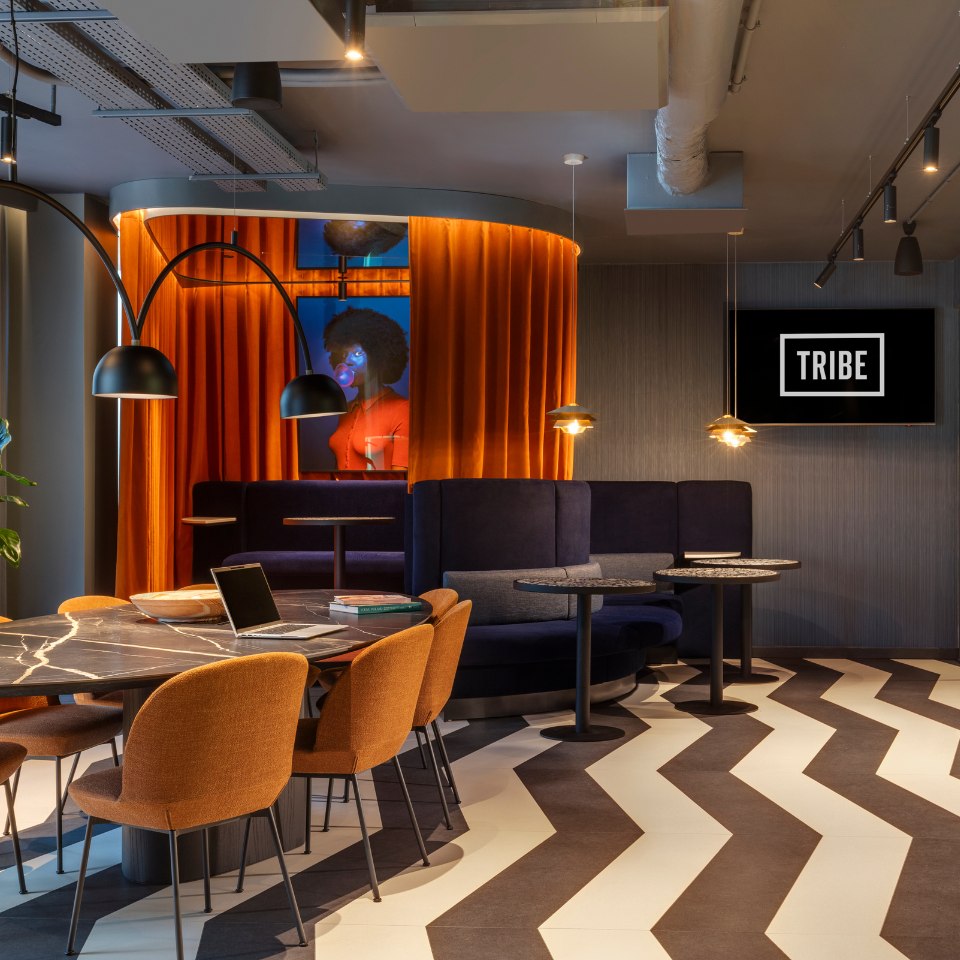 Modern workspace with a curved booth and orange curtains. Dark chairs surround a marble table. A TV displays "TRIBE." The ambiance is sleek and welcoming.