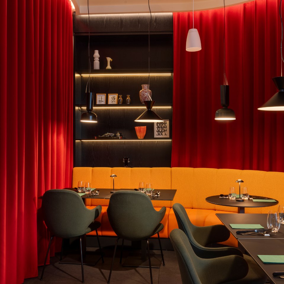 Stylish interior with red curtains, yellow booth seating, green chairs, and black tables. Warm lighting and decorative shelves enhance the cozy ambiance.