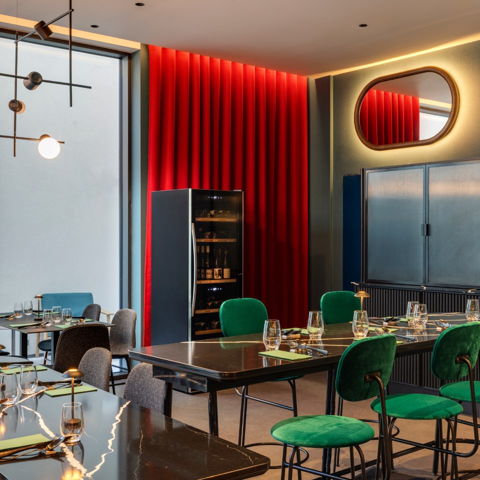 Modern restaurant with green chairs, black tables, and gold accents. Red curtain backdrop and sleek lighting create a chic, elegant atmosphere.