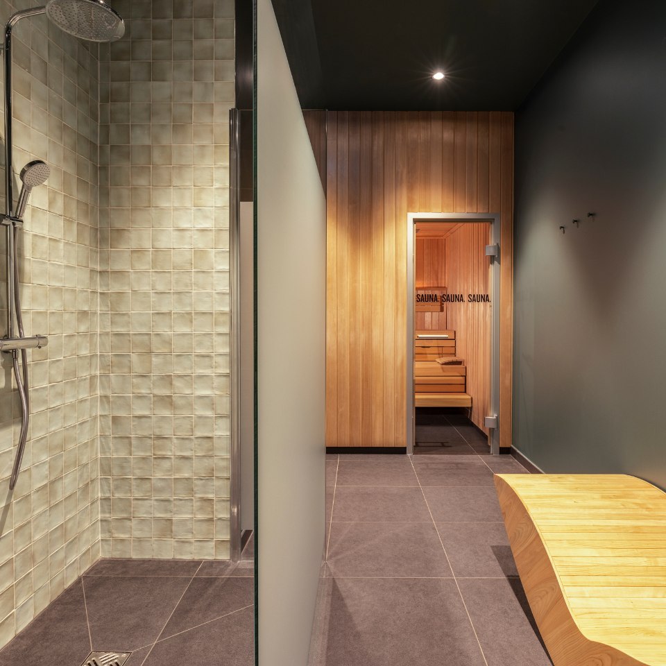 Modern shower and sauna room with sleek design. The shower has textured tiles and glass partition; a warm, inviting wooden sauna is seen in the background.
