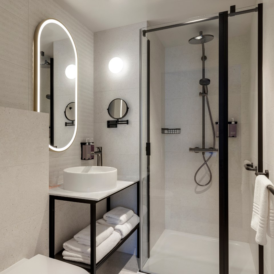 Modern bathroom with sleek design. Features glass shower, oval mirror with LED lighting, round sink on a black metal stand, and folded towels below.