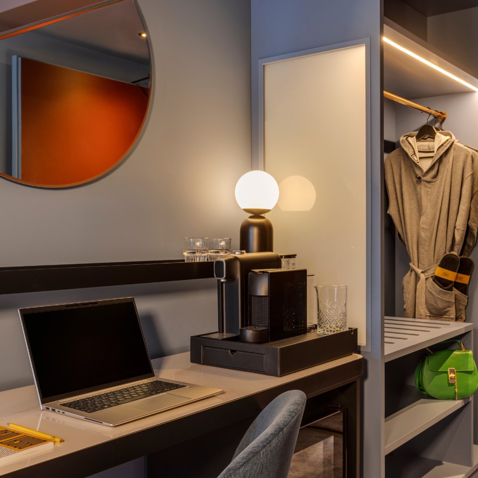 Modern hotel room desk with a laptop, coffee maker, and glowing lamp. A round mirror reflects a warm light. Robe and green handbag in open closet.