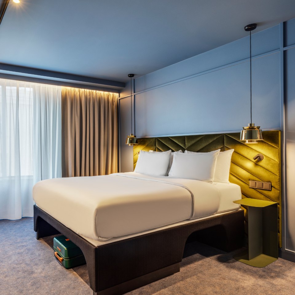 A modern hotel room with a neatly made bed featuring white linens and cushions. The green velvet headboard contrasts with the soft blue walls, creating a calm ambiance. Pendant lights hang symmetrically, illuminating the space elegantly.