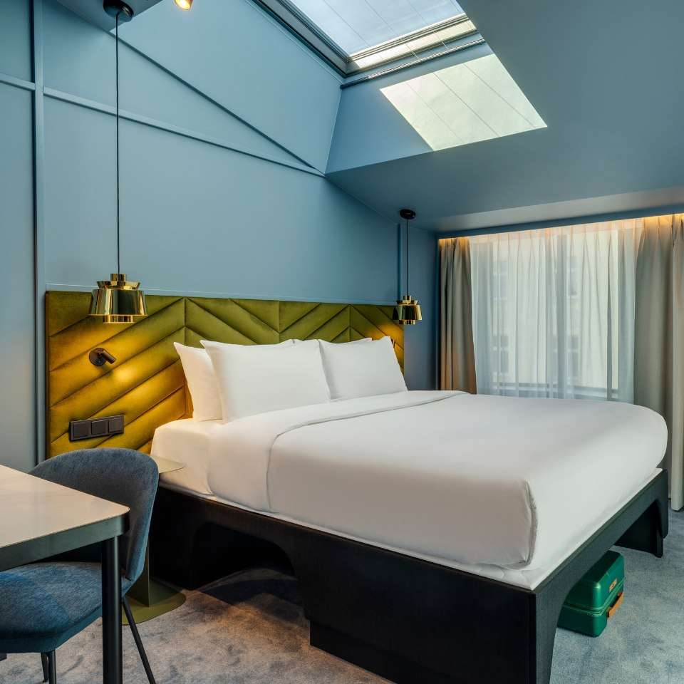 Modern hotel room with a large bed, white linens, and a green cushioned headboard. Skylight above and soft lighting create a cozy ambiance.