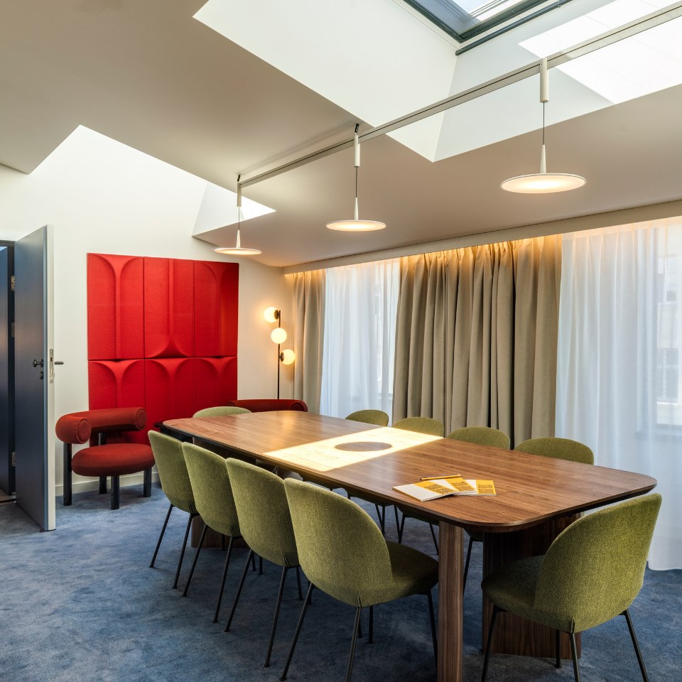 Modern conference room with a long wooden table, surrounded by green chairs on a blue carpet. Red wall panels and soft lighting create a warm atmosphere.