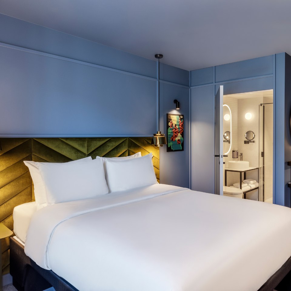 Modern hotel room with a large bed featuring white linens, a green velvet headboard, and blue walls. A door opens to a well-lit, stylish bathroom.