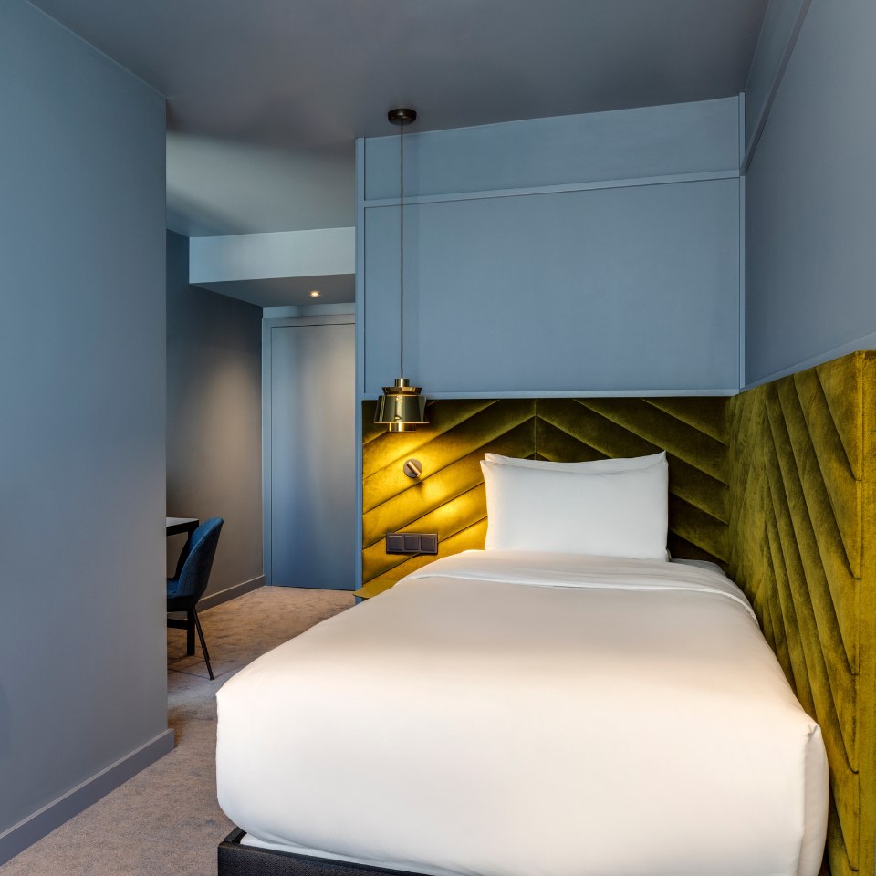 A cozy hotel room with modern design features a single bed with a white duvet. The bed is against an olive-green textured accent wall. A black pendant lamp hangs above, and a small desk with a chair is in the corner. The room's soft lighting and muted blue-gray walls create a calm, inviting atmosphere.