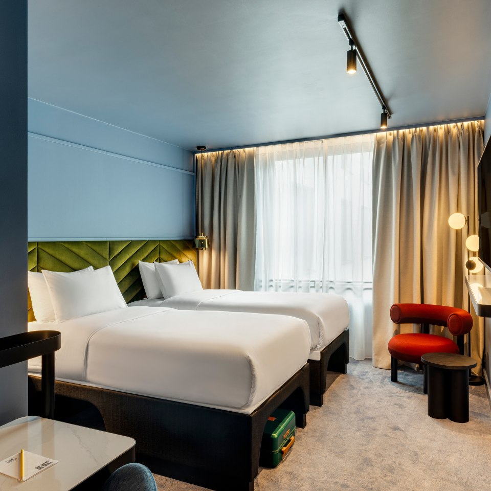 Modern hotel room with two neatly made beds, light blue walls, and a stylish green headboard. Soft natural light filters through sheer curtains. A red chair and minimalist lighting add elegance.