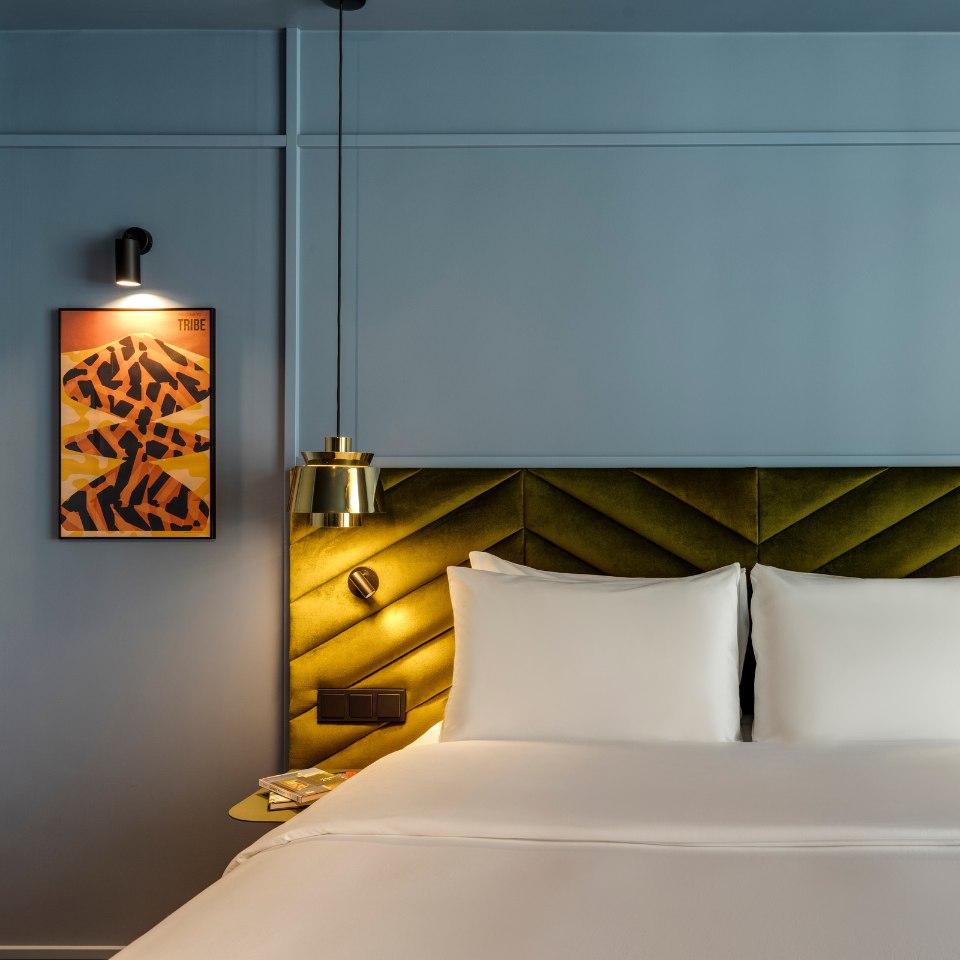 A modern hotel room features a bed with crisp white linens and an olive-green, chevron-patterned headboard. A sleek gold pendant light hangs above, while a framed abstract artwork with warm tones is illuminated by a wall lamp, creating a cozy and sophisticated ambiance.