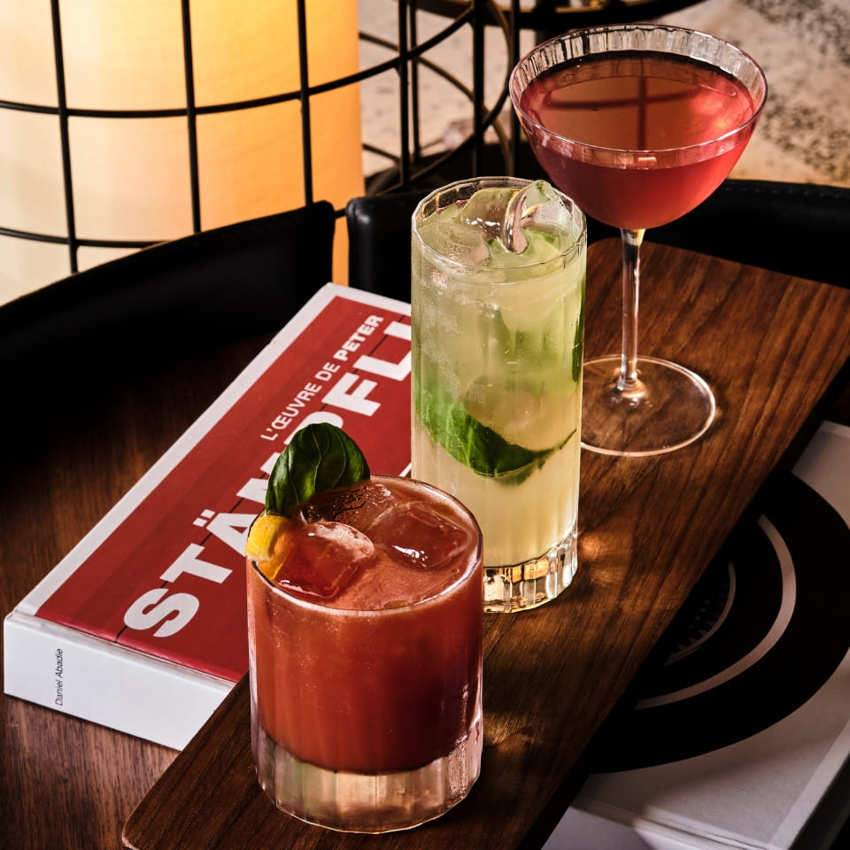 Three elegant cocktails sit on a wooden tray: a red one with basil, a tall green mint drink, and a pink martini. A book and warm light add a cozy vibe.