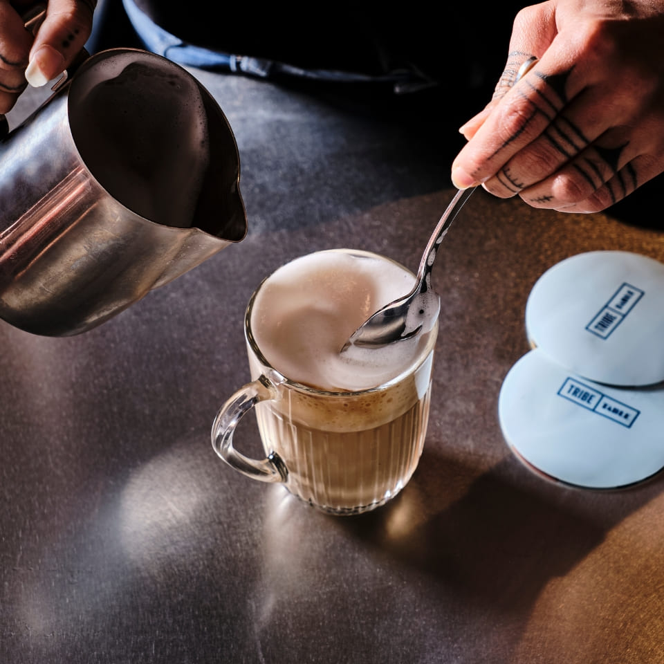 A hand with tattoos gently spoons foam onto a frothy latte in a glass mug. A metal pitcher and two blue coasters are on the shiny surface. Cozy and artisanal.