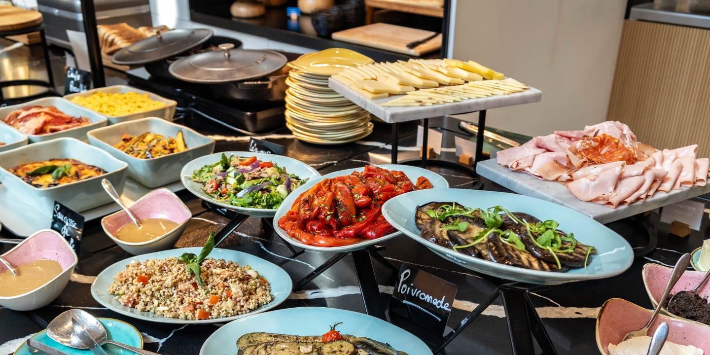 Buffet table with an array of dishes, including roasted peppers, grilled eggplant, mixed salad, cold cuts, cheese, and quinoa salad, creating a vibrant and appetizing display.