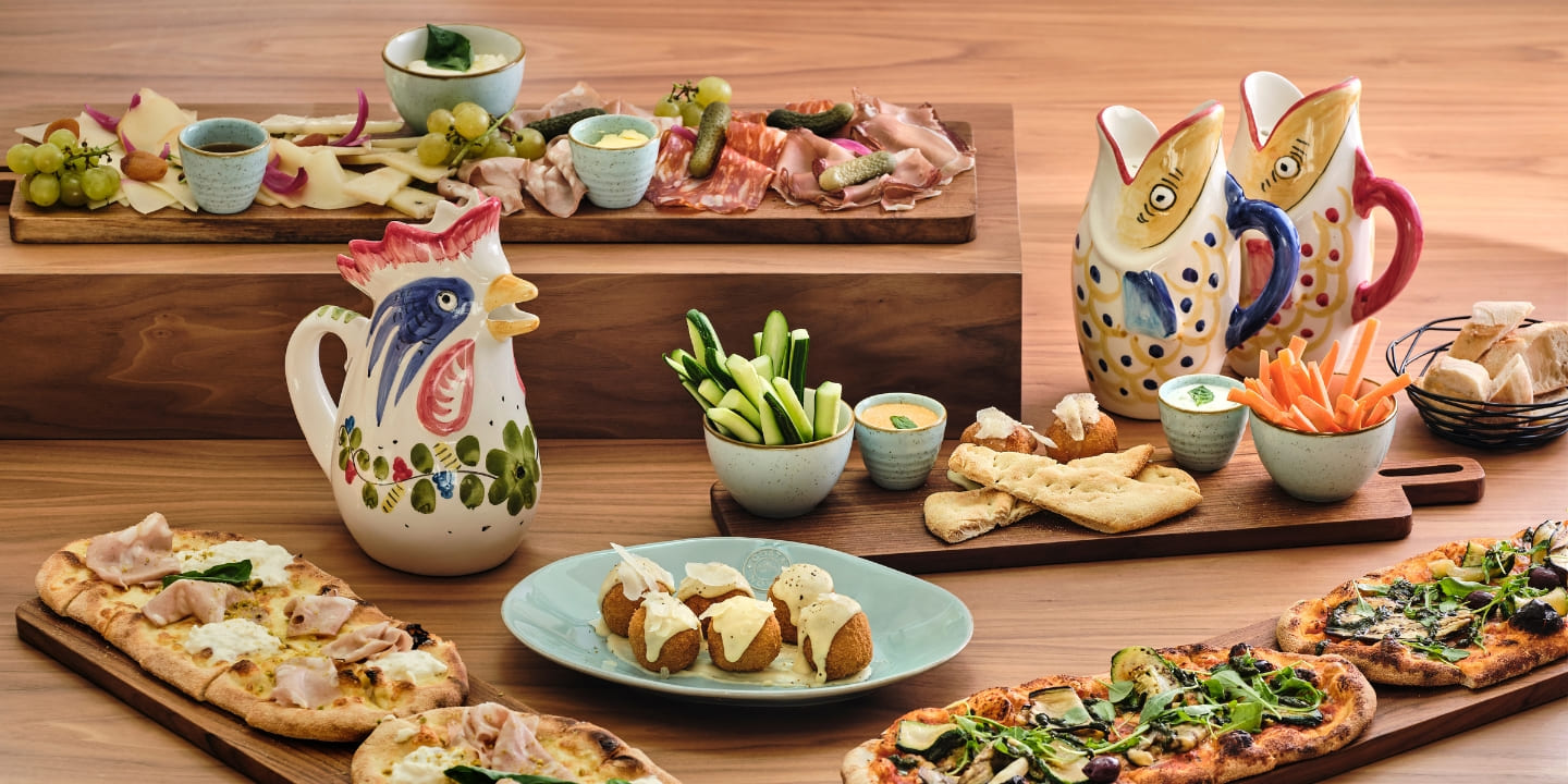 A rustic table with charcuterie boards, flatbreads, dips, and ceramic rooster pitchers. The arrangement is colorful, inviting, and appetizing.