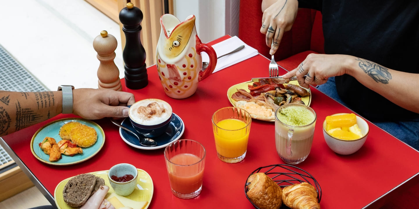 Colorful brunch spread on a red table with two people. Plates of hash browns, bacon, and sausages, plus drinks, a cat-shaped pitcher, and pastries, create a lively, appetizing scene.
