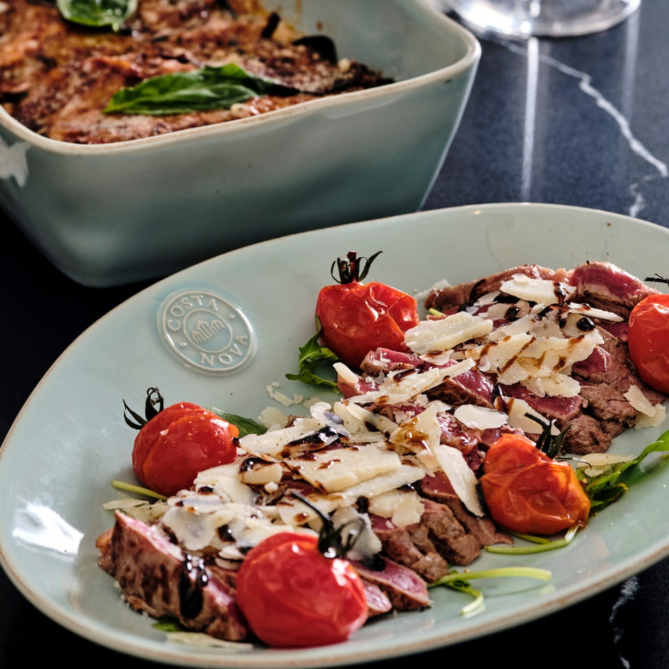 A plate of sliced steak topped with shaved cheese and balsamic glaze, garnished with roasted cherry tomatoes and greens. A dish of baked pasta in the background.