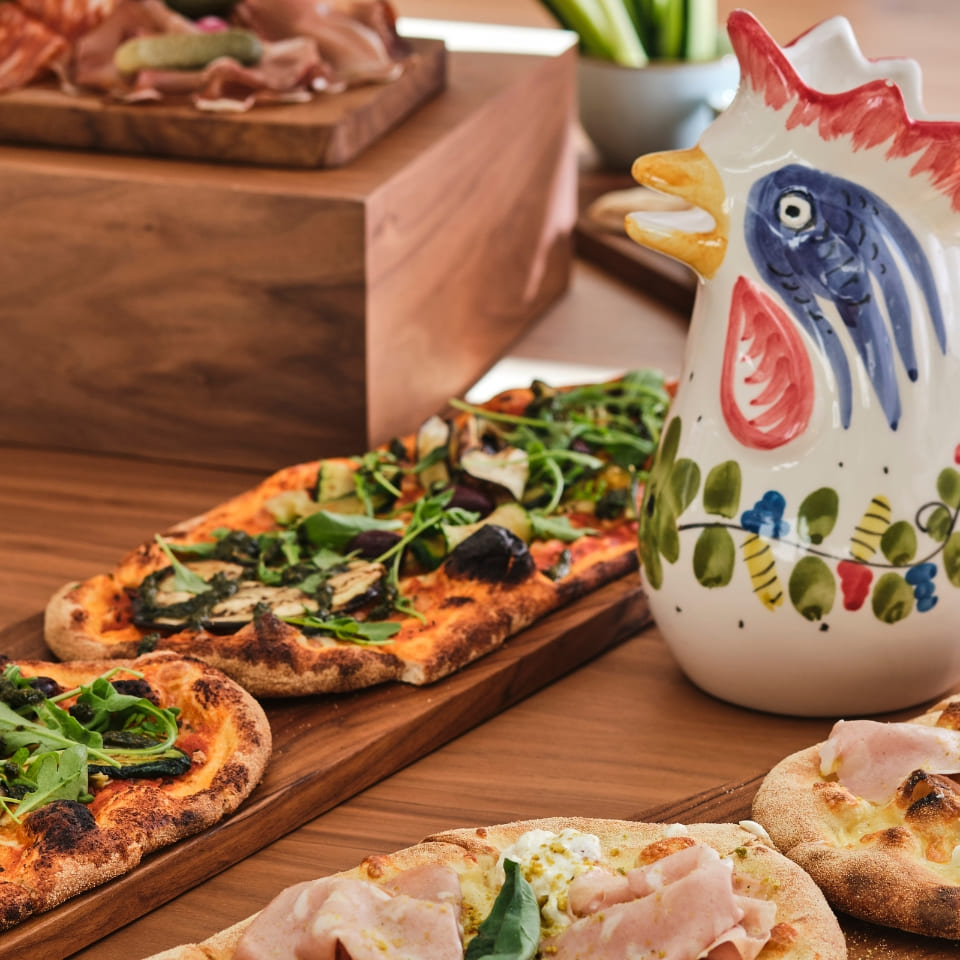 A vibrant ceramic rooster pitcher sits beside wooden trays of flatbread topped with greens and meats, creating a warm, rustic dining atmosphere.