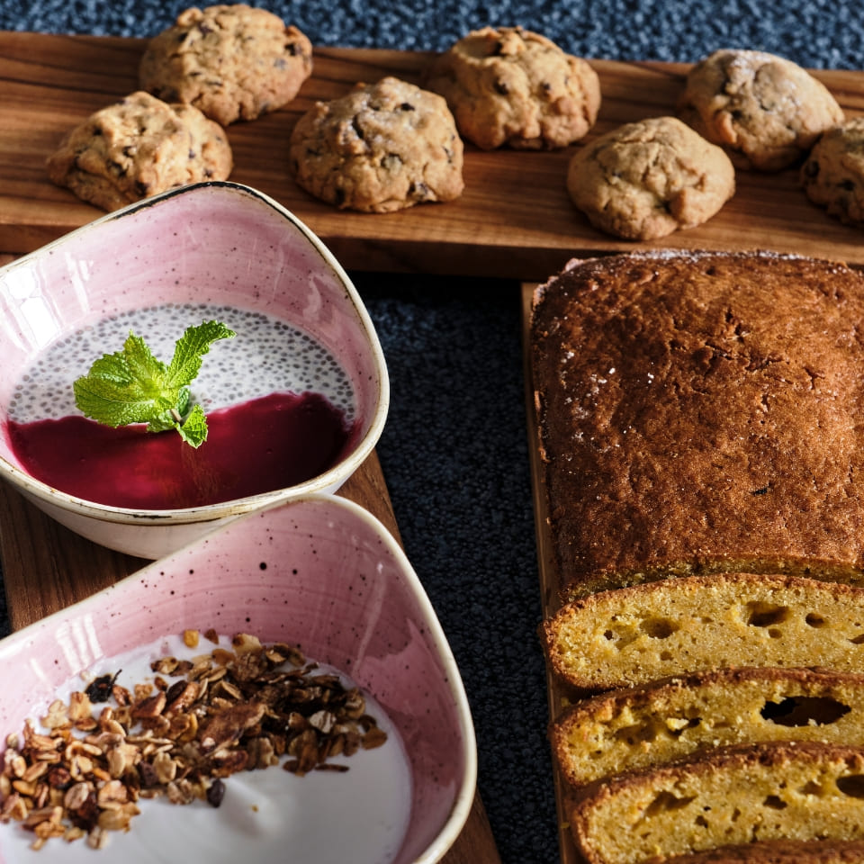 A cozy breakfast spread with chia seed pudding topped with mint, walnut bread, cookies, and yogurt with granola, on a textured blue surface.