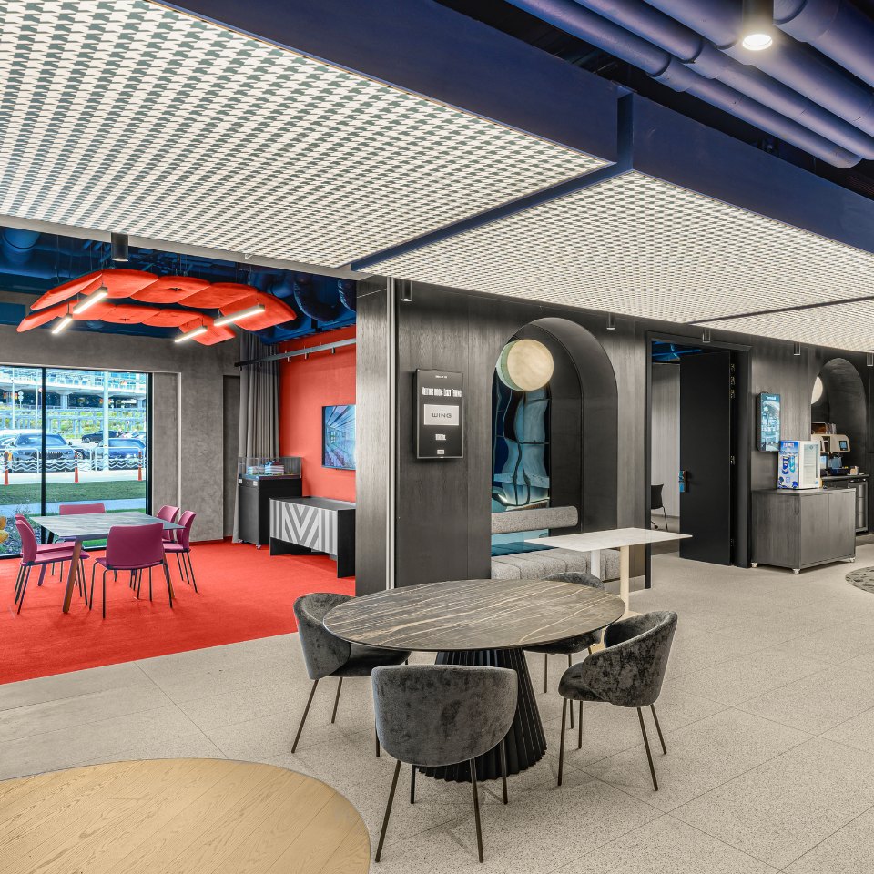 Modern office lounge with a sleek design featuring gray and red tones. Round tables, velvet chairs, and large windows create an open, inviting atmosphere.