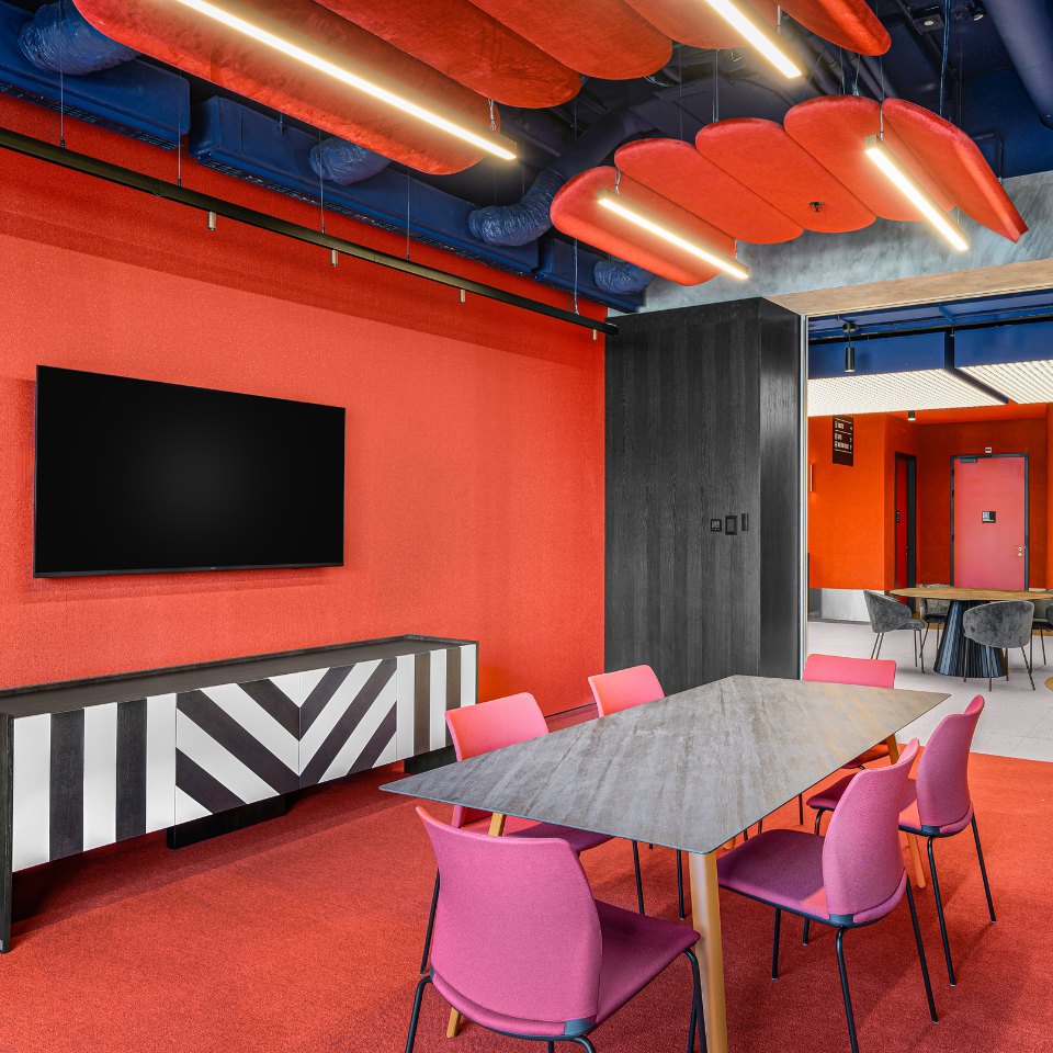 Modern meeting room with bold red walls and ceiling accents. Features a triangular gray table, pink chairs, striped cabinet, and a wall-mounted TV.