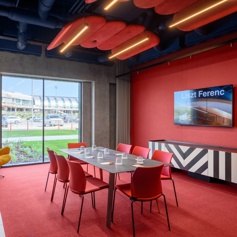 Modern meeting room with red chairs and table, vibrant red walls, large window, and ceiling lights. A screen displays "Liszt Ferenc." Spacious and stylish.