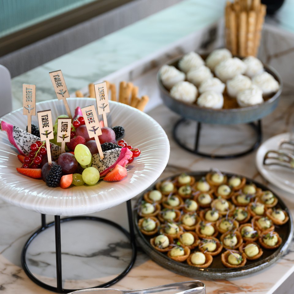 A table with a white plate of fresh fruit, including dragon fruit and grapes, with wooden picks labeled "Libre." Nearby are trays of pastries and breadsticks.