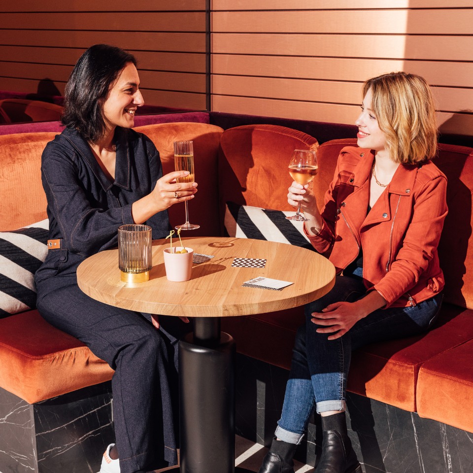 Two women sit at a cozy café. They're smiling, toasting with drinks, creating a warm and friendly atmosphere. The setting feels modern and inviting.