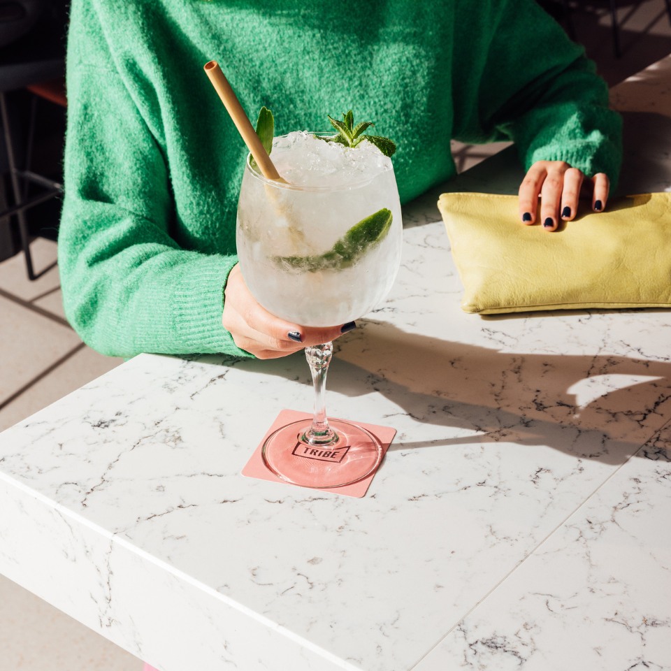 A person in a green sweater holds a cocktail with lime and mint on a marble table. A yellow clutch rests nearby, creating a fresh, vibrant atmosphere.