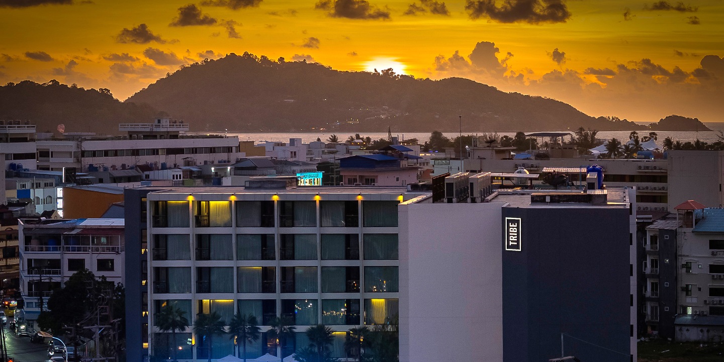 Sunset over cityscape with illuminated Tribe hotel building, palm trees, and distant hills against a vibrant orange sky.