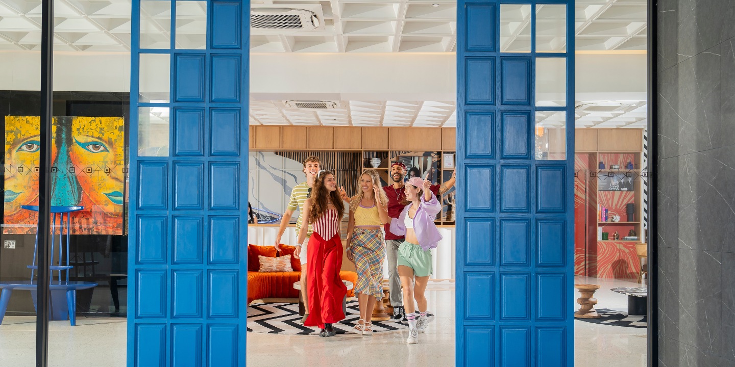 Group of young adults walking through a modern blue doorway into a bright lounge area with orange sofa and patterned rug.