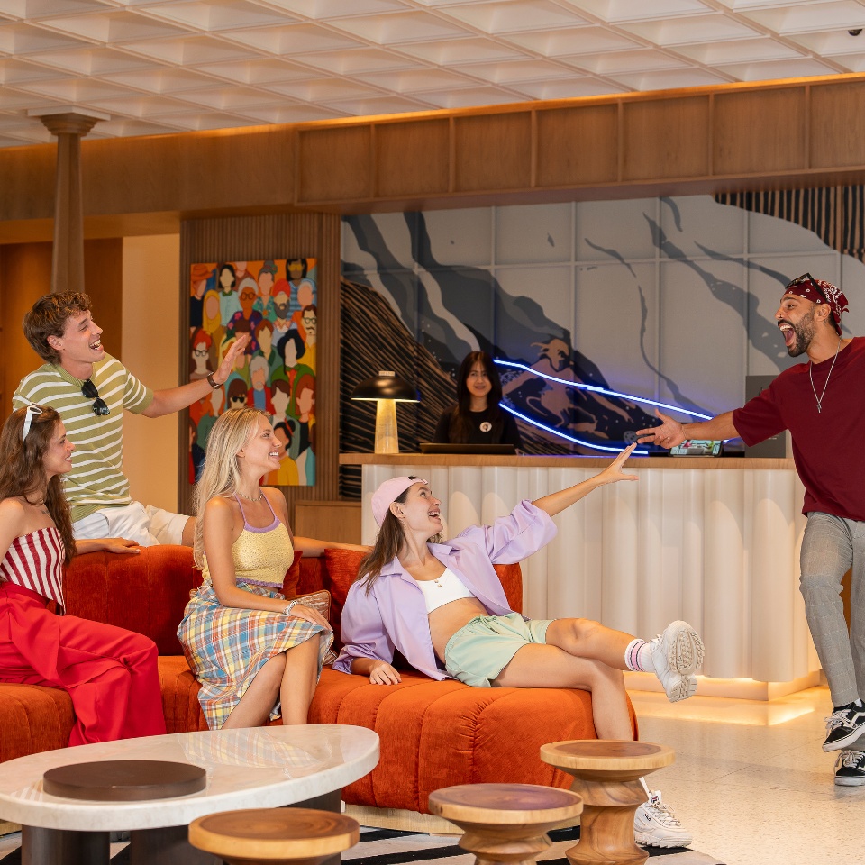 Group of casually dressed young adults interacting in a modern, colorful lounge area with bright orange seating and artistic wall decor.