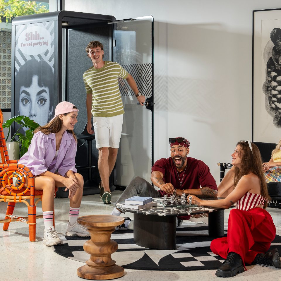 Group of young adults hanging out in a modern room, playing chess and sitting near a closed phone booth with "Shh... I'm not partying" sign.