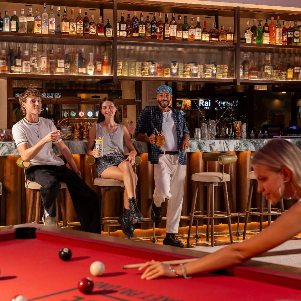 Four people socializing at a bar with drinks, with one person playing pool in the foreground. Bottles of alcohol displayed behind the bar.