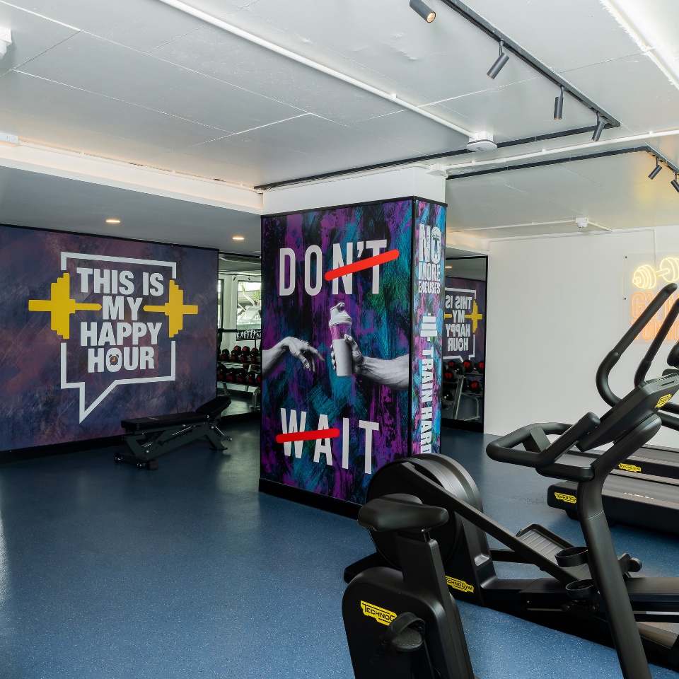 Modern gym interior with motivational murals. A wall says "THIS IS MY HAPPY HOUR" with a weight graphic. Equipment includes treadmills and weights, creating an energetic and inspiring atmosphere.