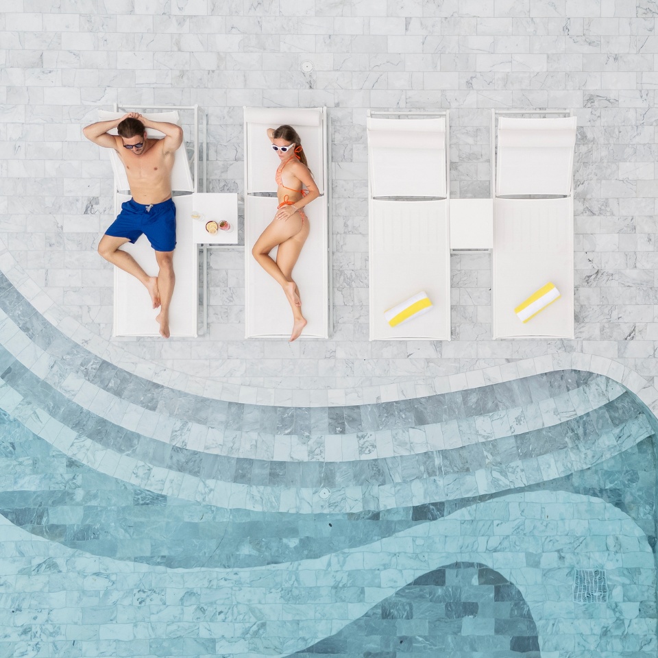 Aerial view of a man in blue shorts and woman in orange bikini sunbathing on loungers beside a curving pool. Bright marble tiles and a relaxed vibe.
