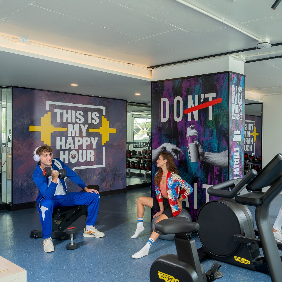 A gym scene with a man in a blue tracksuit lifting dumbbells and a woman in colorful attire stretching. Motivational phrases decorate the walls.
