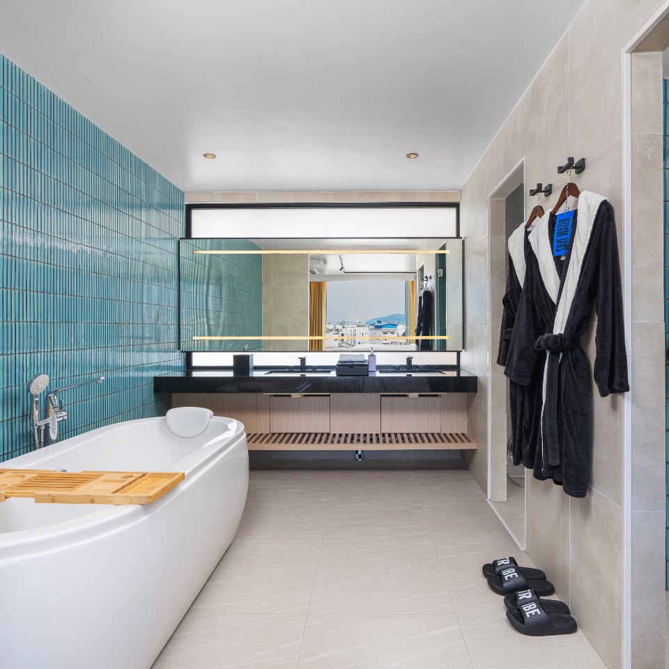 Modern bathroom with a white bathtub, teal tiled wall, double sink vanity, large mirror, black robes, and matching slippers.